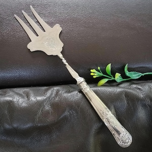 May include: A silver-toned metal serving fork featuring a three-pronged head with an engraved floral pattern. The neck of the fork has a twisted design, and the handle is decorated with embossed scrollwork and a shell motif at the base.