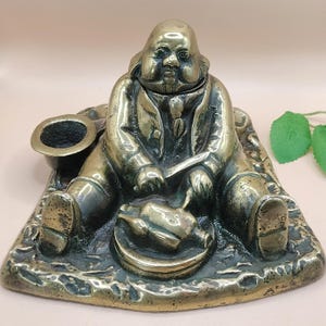 May include: A bronze-colored figurine of a seated figure, possibly Benjamin Franklin, holding a knife and a plate with a cooked bird. A hat sits nearby. The figurine is on a textured base.