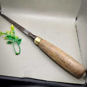 May include: A woodworking chisel with a wooden handle and a metal blade. The handle is light brown with a brass ferrule. The blade is dark gray and tapers to a point. A small green and yellow plant is in the background.