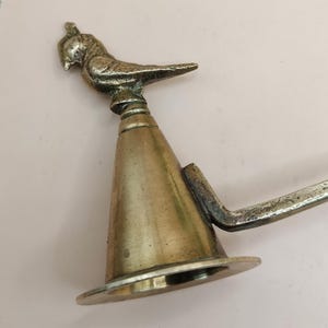 May include: A vintage brass candle snuffer with a bird-shaped handle. The snuffer has a conical body and a circular base. The bird is perched atop the cone, adding a decorative element to the functional piece. The metal has a tarnished, antique finish.