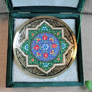 May include: A decorative, round, gold-toned plate with an intricate floral design, featuring red pomegranates and blue and white flowers on a green background. The plate is presented in a green velvet box with a white patterned lining.