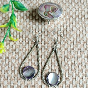 May include: A pair of silver-toned drop earrings featuring a twisted metal frame and a circular iridescent abalone shell inlay, accompanied by a matching oval-shaped brooch with a swirling purple, green, and white pearlescent shell surface.