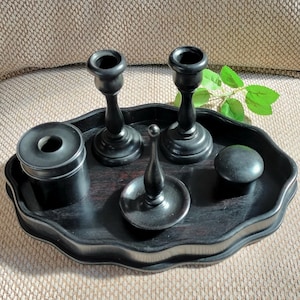 Könnte beinhalten: Ein schwarzes Holz-Waschtisch-Set auf einem Tablett mit gewelltem Rand. Das Set enthält zwei Kerzenhalter, einen kleinen runden Behälter, eine kleine Schale mit dekorativer Oberseite und einen runden Knopf. Die Artikel sind auf einer dunklen Holzoberfläche angeordnet.