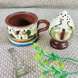 May include: A ceramic mug with a brown interior and handle, featuring a landscape scene with the word "Poole." A matching salt shaker and a small metal shovel are also present. The mug has a blue and brown base.