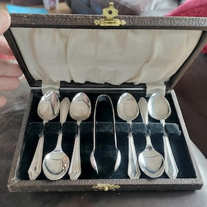 May include: A set of silver spoons and tongs displayed in a brown, hinged box with a cream-colored satin lining. The spoons have a classic design, and the box is closed with a gold-colored clasp.