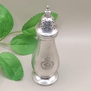 Könnte beinhalten: Ein silberfarbener Salzstreuer mit rundem Boden und perforierter Oberseite. Der Salzstreuer hat ein dekoratives Emblem mit der Aufschrift "Army Canteen Board India". Das Design ist vintage und der Salzstreuer ist wahrscheinlich aus Metall.