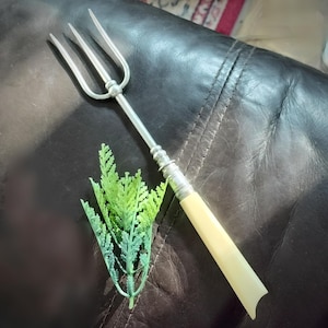May include: A silver-toned metal serving fork featuring three long tines and a smooth, cream-coloured handle. The handle has a cylindrical shape with a slightly flared end, and the metal shaft connects to the tines with a small decorative sphere.