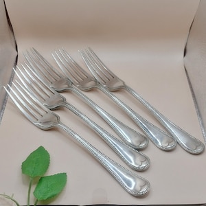 May include: Six silver forks with a decorative beaded edge and a heart-shaped design at the base of the handle. The tines are long and slender. The forks are arranged on a light beige surface.