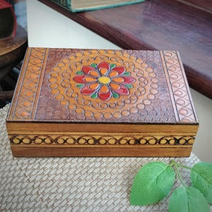 May include: A wooden box with a carved floral design on the lid. The flower is painted in shades of red, orange, yellow, and green. The box has a brown wood finish and a decorative border around the edge.