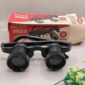 May include: Black spectacles-binocular with a red and brown box. The box has the words "SPECTACLES-BINOCULAR" printed on it. The spectacles-binocular has the number "504" printed on the box and on a small tag.