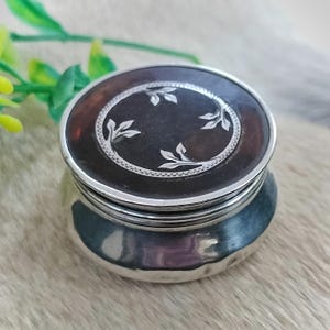 May include: A small, round silver container featuring a polished metallic base and a dark brown, circular lid. The lid is decorated with a silver-toned circular border and a delicate, engraved leaf pattern centered on the dark surface.