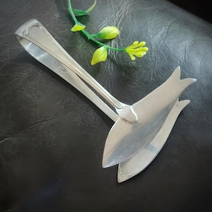 May include: A pair of silver-toned metal sugar tongs featuring a polished finish and a distinctive fish-shaped design at the gripping ends. The tongs have a long, slender handle that curves into a spring-loaded mechanism, with a hallmark visible.