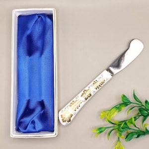 May include: A butter knife with a white handle decorated with gold floral patterns. The stainless steel blade is next to a white box with a blue satin lining. The knife is approximately 7 inches long.