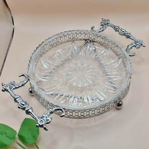 May include: A clear glass serving dish with a starburst pattern in the centre, set within a silver-coloured metal filigree frame. The dish has two ornate handles, ideal for serving appetizers or desserts.