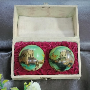May include: Two spherical meditation balls with green surfaces featuring painted illustrations of a stone bridge and landscape, resting inside a rectangular box lined with deep red velvet and a cream-coloured fabric lid interior.