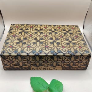 May include: A rectangular wooden box with a floral pattern carved into the lid. The flowers are cream-colored with red and black accents. The word "KRYNICA" is carved on the front of the box.
