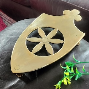 May include: A polished brass trivet shaped like a shield with a pointed bottom and a decorative top finial. The center features a cutout design of a six-petaled flower. The metal surface is smooth and reflective with a warm golden yellow tone.