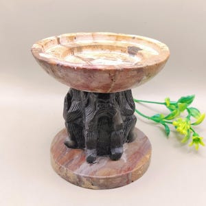 May include: A decorative stone bowl with a carved base. The bowl is a light brown color with a circular design. The base is dark gray and features a carved design. The base sits on a round, reddish-brown base.