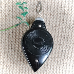 May include: A black Stanley Chalk-O-Matic chalk line tool with a teardrop-shaped plastic casing, a silver metal hook attached to a string, and a textured surface. The centre of the tool features circular grooves and the text Made in U.S.A.