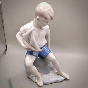 May include: A porcelain figurine of a young person in a white shirt and blue shorts, holding a small sailboat. The figure is seated on a light blue rock-like base, with a serene expression. The figurine is a decorative item.