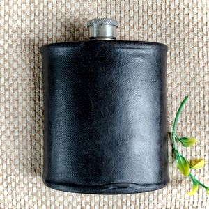 Könnte beinhalten: Ein rechteckiger schwarzer Flachmann mit einer glatten, dunkel lederartigen Oberfläche und einem silberfarbenen Metallschraubverschluss. Der Flachmann hat abgerundete Ecken und einen leicht gewölbten Körper, was ein schlichtes Design zur Aufbewahrung von Flüssigkeiten ergibt.