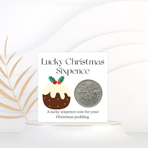 Op de afbeelding: Een witte kaart met de tekst "Lucky Christmas Sixpence" toont een cartoon kerstpudding en een zilveren munt. De tekst hieronder luidt "Een geluksmunt van zes pence voor je kerstpudding."