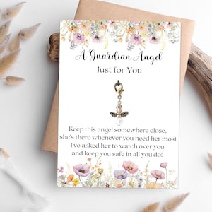 Könnte beinhalten: Eine weiße Karte mit floralen Akzenten und dem Text "A Guardian Angel Just for You". Ein Anhänger in Form eines Engels ist an der Karte befestigt. Die Karte liegt auf einem braunen Umschlag.