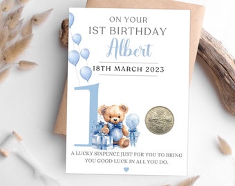 May include: A blue and white card with a teddy bear holding a gift and a blue balloon. The card says "On your 1st birthday Albert 18th March 2023" and "A lucky sixpence just for you to bring you good luck in all you do."