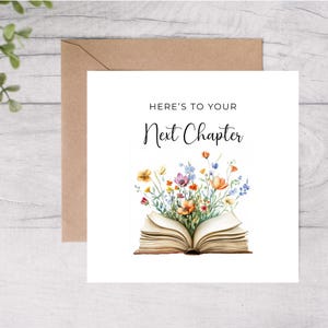 Könnte beinhalten: Eine Grußkarte mit dem Text "Here's to your Next Chapter" in schwarzer Schrift. Die Karte zeigt ein offenes Buch mit bunten Aquarellblumen, die aus den Seiten quellen. Ein brauner Umschlag befindet sich hinter der Karte.