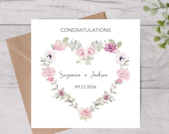May include: A white wedding congratulations card featuring a floral heart design. The heart is made of pink and purple flowers and green leaves. The names "Suzanna" and "Jackson" are written inside the heart, along with the date 09.12.2026. The card is on a brown envelope.