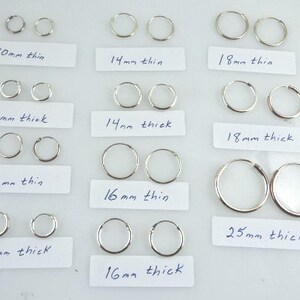 May include: A collection of silver hoop earrings in various sizes and thicknesses. The earrings are displayed on white labels with handwritten measurements in millimeters, including 10mm, 12mm, 14mm, 16mm, 18mm, and 25mm.