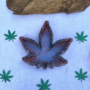 May include: A purple and red glitter resin ashtray shaped like a marijuana leaf. The ashtray is sitting on a white surface with green marijuana leaf cutouts.
