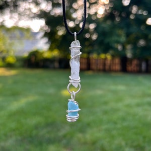 Op de afbeelding: Een handgemaakte hangerketting met een heldere kwartskristal gewikkeld in zilverdraad, hangend aan een zwart koord. Daaronder is een kleine, lichtblauwe kristal ook in zilverdraad gewikkeld. De achtergrond is een vage buitenscène.