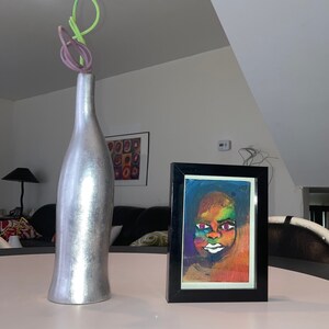 May include: A silver vase with a green and purple stem on a white table next to a framed painting of a colourful portrait of a person with a white background.