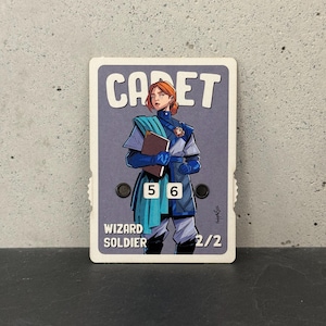 May include: A rectangular game card featuring an illustration of a red-haired character in a blue uniform holding a book. The card displays the text CADET, WIZARD SOLDIER, and 2/2. Two black circular dials are integrated into the card, showing the numbers 5 and 6.