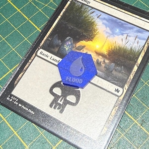 May include: A Magic: The Gathering card labeled "Swamp" with a blue, hexagonal token that reads "Flood." The card features a landscape with a sunset and marsh plants. The token is placed over the card's text and skull symbol.