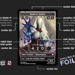 May include: A black card featuring an Eldrazi Spawn creature illustration with blue and white tones. The card includes number dials, a main display, and text indicating it is a token creature. The text "AVAILABLE IN FOIL" is also present.