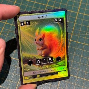 Puede incluir: Una carta de comercio de lámina verde, dorada e iridiscente con una imagen de una ardilla de dibujos animados. La carta tiene el texto "Squirrel" y "Token Creature - Squirrel" impreso en ella. La carta tiene una fuerza y resistencia de 1/1.