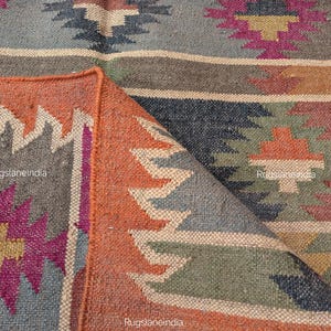 Peut inclure: Un tapis tissé multicolore avec un motif géométrique. Le tapis présente des nuances d'orange, de gris, de marron, de vert et de violet. Le bord du tapis est replié, révélant le dessous à franges. Idéal pour la décoration intérieure.