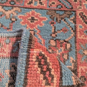 Puede incluir: Primer plano de una sección de una alfombra con un patrón geométrico detallado. La alfombra presenta una combinación de colores, incluyendo tonos de azul, rosa y marrón. El diseño incorpora formas florales y geométricas, con un borde doblado que revela el grosor de la alfombra.