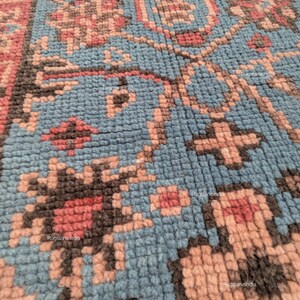 Puede incluir: Primer plano de una sección de una alfombra con un patrón detallado y texturizado. La alfombra presenta un fondo azul claro con diseños florales y geométricos en tonos rosa, marrón y negro. El diseño es intrincado y parece estar anudado a mano.