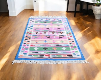 Könnte beinhalten: Ein bunter Teppich mit einem geometrischen Muster in Rosa-, Blau-, Grün- und Weißtönen. Der Teppich hat einen Fransenrand und liegt auf einem Holzboden.