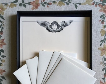 May include: A black box with a set of white stationery featuring an angel with wings and a face at the top of the page.