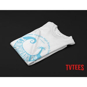 May include: White t-shirt with a light blue graphic design. The design includes the text "Tsunami Drama 06" in a curved font, along with "Summer Heights High Drama Company presents" and a stylized palm tree.