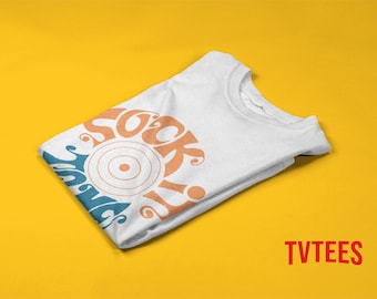 Könnte beinhalten: Weißes T-Shirt gefaltet auf gelbem Hintergrund. Das Shirt zeigt den Text "Sock It" in Orange und Blau mit einer sonnenartigen Grafik. Der Markenname "TVTEES" ist rot gedruckt.