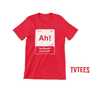 May include: A bright red short-sleeved t-shirt featuring a white rectangular graphic designed like a periodic table element. The graphic includes the text Surprise, 117, Ah!, 14.994, and The Element of Surprise. The word TVTEES appears in red.