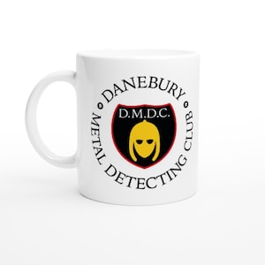 Può includere: Tazza in ceramica bianca con il logo "Danebury Metal Detecting Club". Il logo presenta una figura con elmo giallo su uno scudo nero con bordo rosso. Il testo nero circonda il logo.