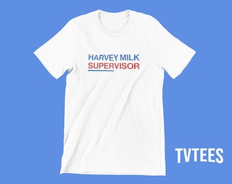 Può includere: T-shirt bianca con la scritta "HARVEY MILK SUPERVISOR" in blu e rosso, con una sottolineatura blu. La maglietta è su uno sfondo blu, con la parola "TVTEES" in bianco nell'angolo in basso a destra.