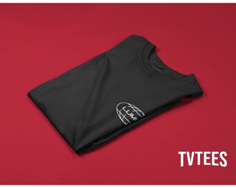May include: A folded black t-shirt with a white logo on the left chest. The logo is a stylized globe with the word "LUMI" inside. The background is a solid red color, with the text "TVTEES" in white.