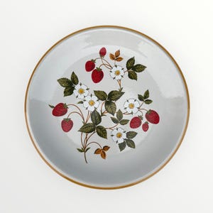 May include: A round, light gray ceramic plate with a brown rim. The plate is decorated with a strawberry plant design featuring red strawberries, white flowers, and green leaves. A charming addition to any kitchen.